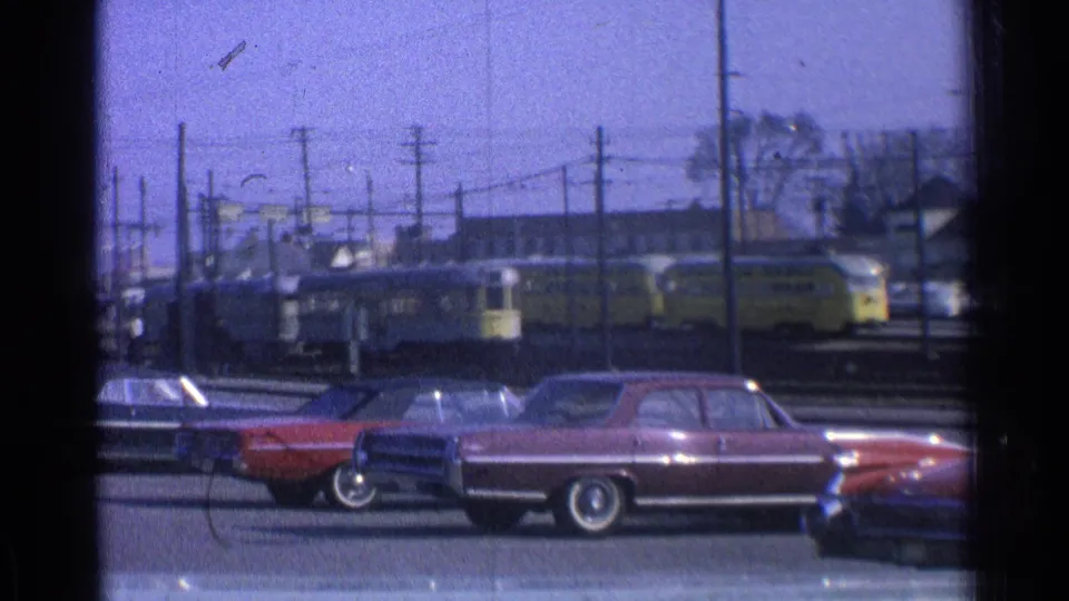 1965 Ohio street view through a dark window frame, with sedans parked before a yellow rail vehicle and overhead wires.