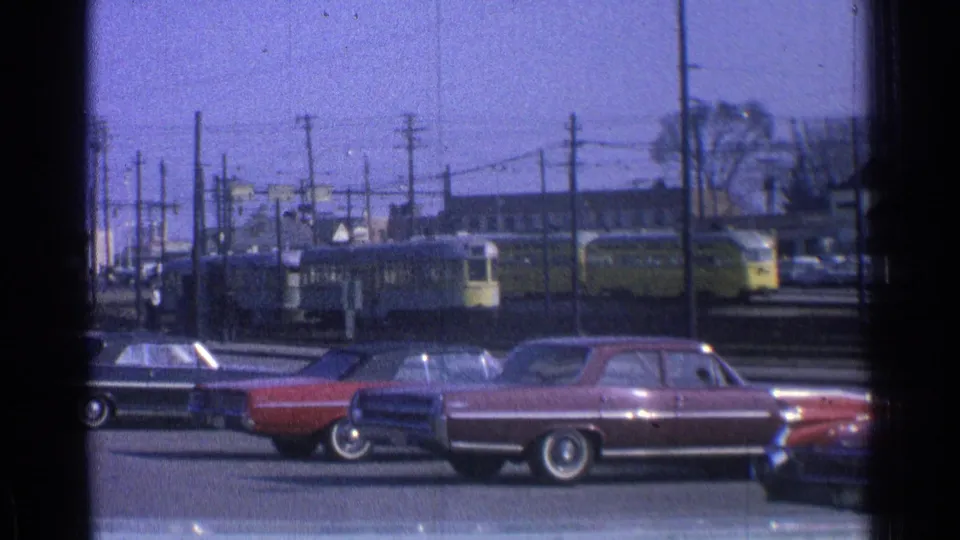 1965 Ohio frame with parked cars in the foreground and a yellow trolley or light rail vehicle beyond the tracks.