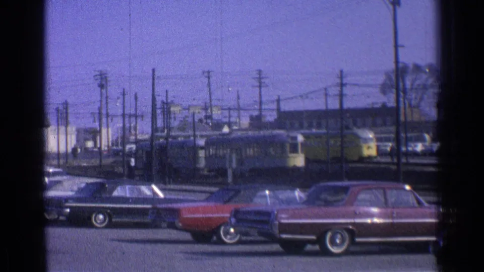 1965 Ohio home-movie frame showing a yellow transit car beyond parked sedans, utility poles, and rail infrastructure.