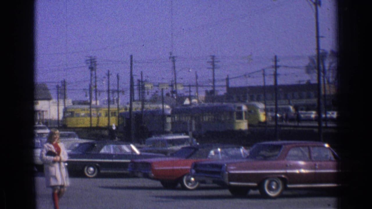 Two women in dress coats cross the foreground of a 1965 Ohio streetcar scene with parked cars and overhead wires.