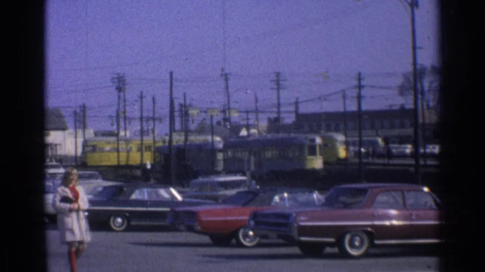 Two women in dress coats cross the foreground of a 1965 Ohio streetcar scene with parked cars and overhead wires.