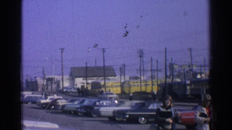 A 1965 Ohio transit view shows yellow streetcars, parked classic cars, overhead lines, and pedestrians entering the foreground.