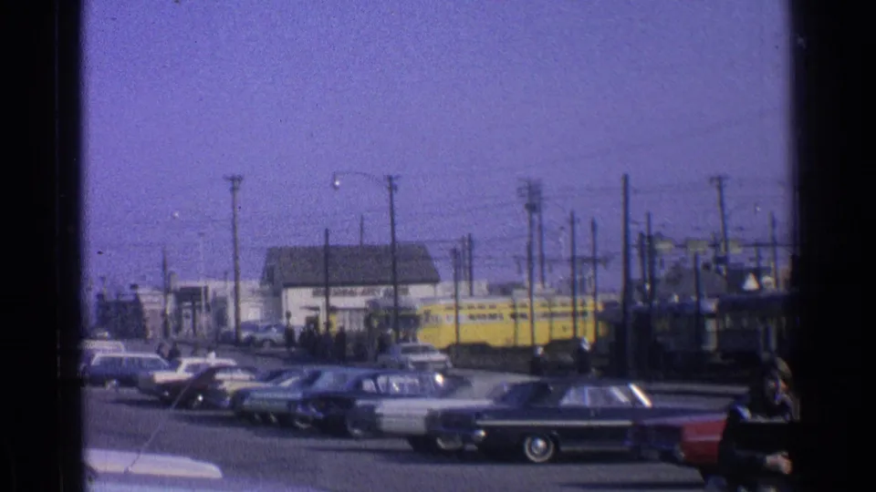 1965 Ohio streetcar scene with a yellow car on tracks, parked automobiles, overhead wires, and a pedestrian at frame edge.