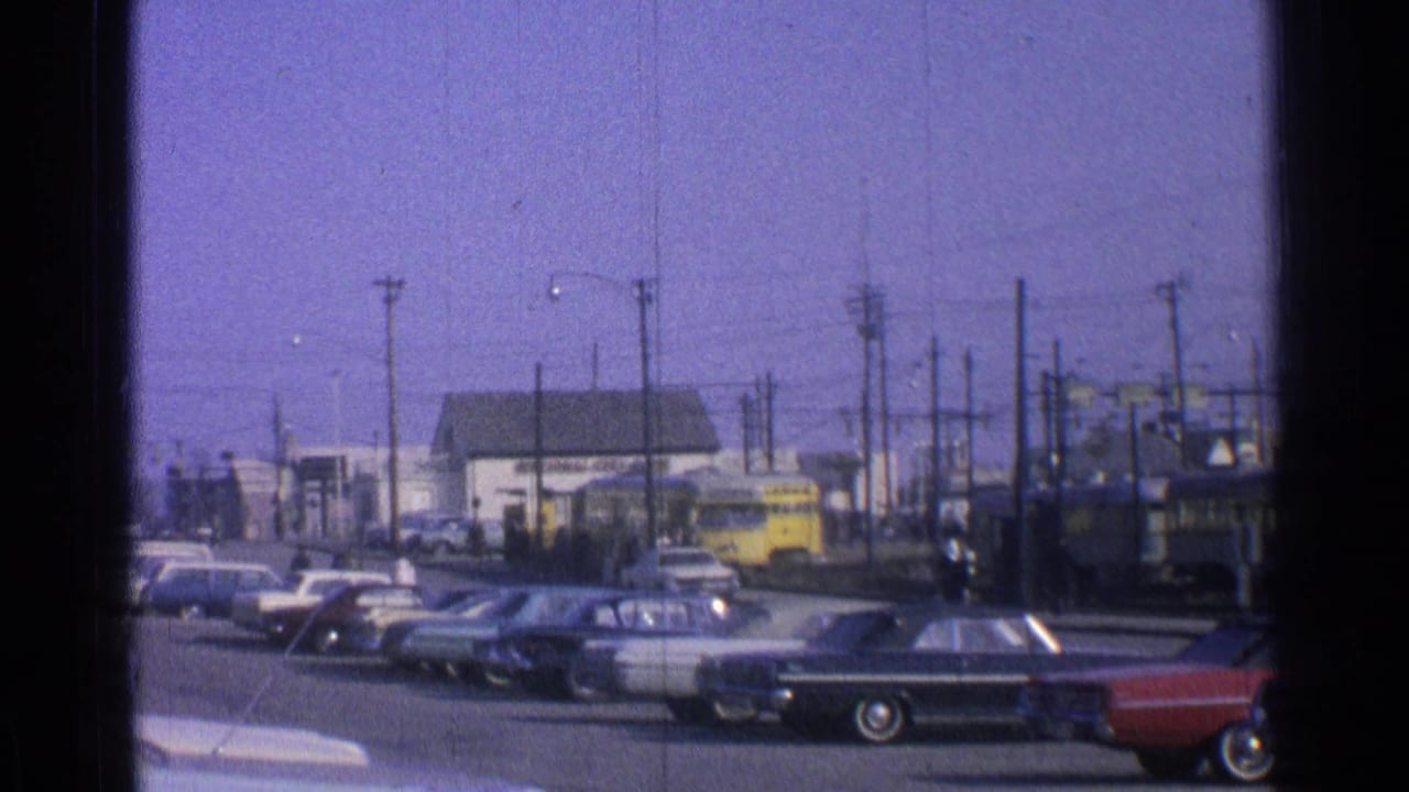 1965 Ohio street view with parked cars in the foreground and a yellow passenger train near a small building behind them.
