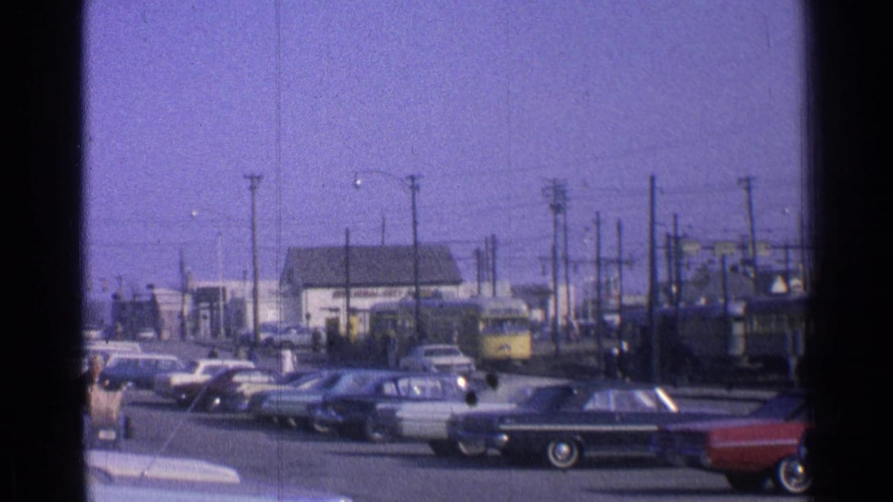 1965 Ohio street scene with classic cars parked along the road while a yellow passenger train moves behind utility poles.