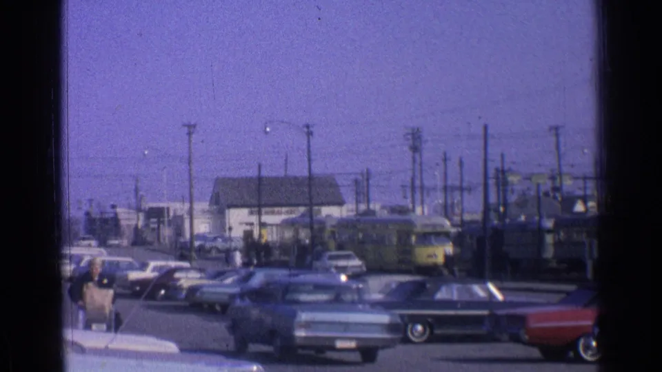 1965 Ohio home-movie frame showing pedestrians near the sidewalk, vintage cars in the street, and a yellow train in the background.