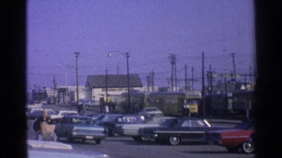 1965 Ohio street view with pedestrians at left, parked midcentury cars, and a yellow passenger train partly visible beyond the road.