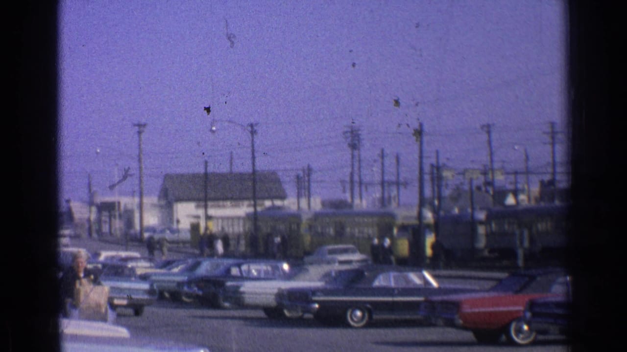 1965 Ohio 8mm street view with parked classic cars, a yellow trolley or rail vehicle, pedestrians and utility poles.