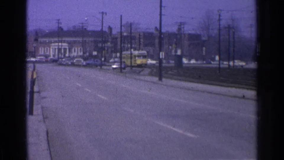 1965 Ohio 8mm road view with brick buildings, utility poles, parked cars and rail or trolley infrastructure along the street.