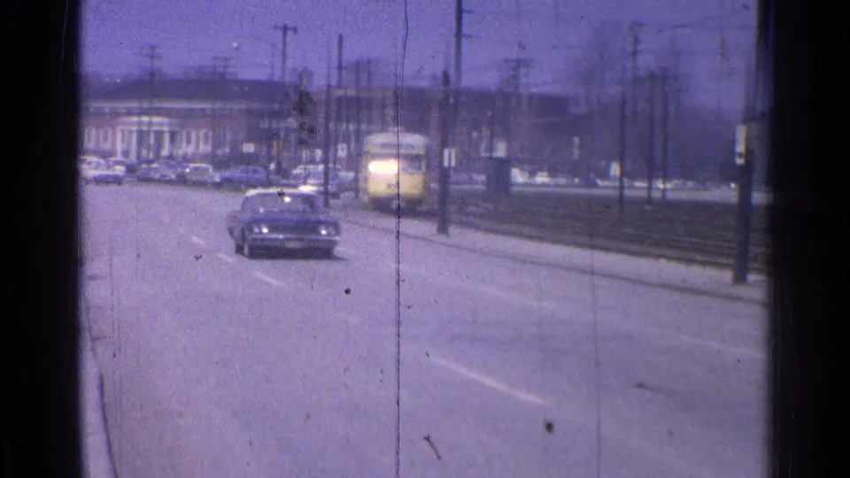 1965 Ohio street view with a car approaching the camera, utility poles, brick buildings and a yellow trolley or rail vehicle behind it.
