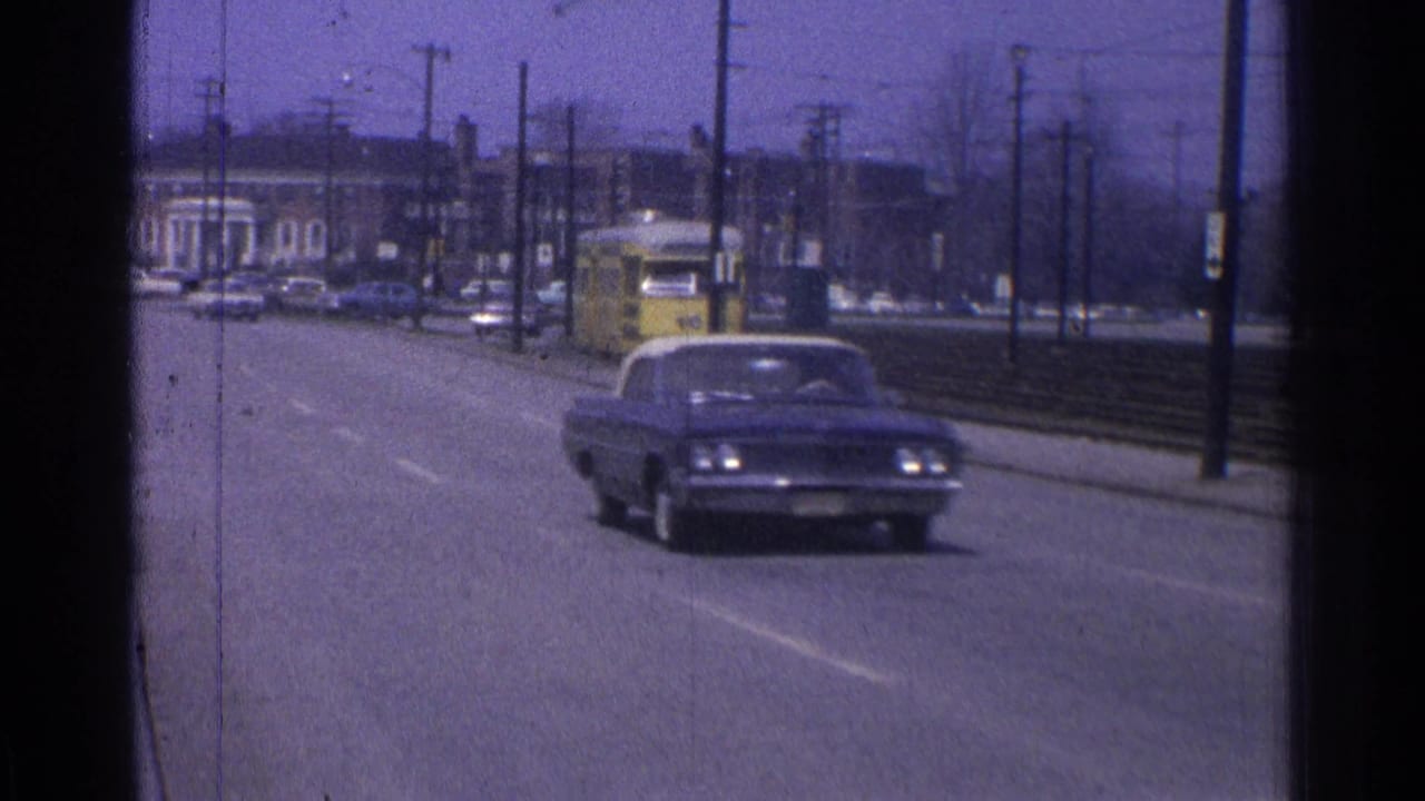 A dark car drives on a broad road near trolley tracks and utility poles.