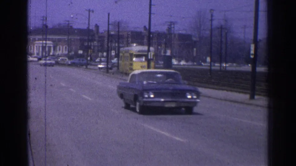 1965 Ohio home-movie frame showing a dark mid-century car on a wide road beside trolley tracks and distant buildings.