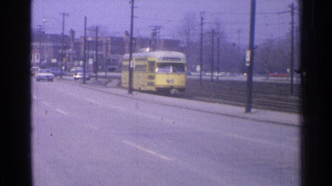 A yellow trolley car travels beside a road with tracks and utility poles.