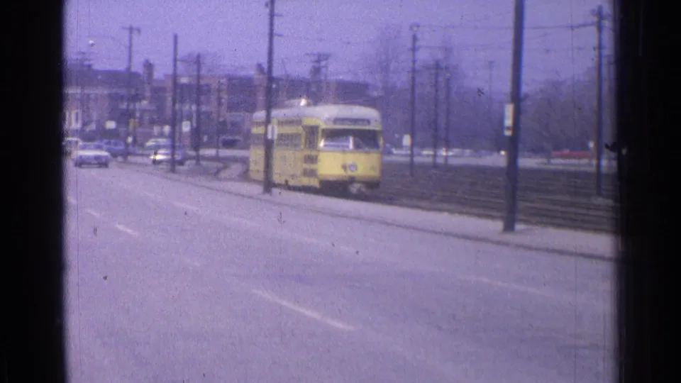 1965 Ohio frame with a yellow trolley moving along rails beside a mostly open roadway and mid-century street infrastructure.