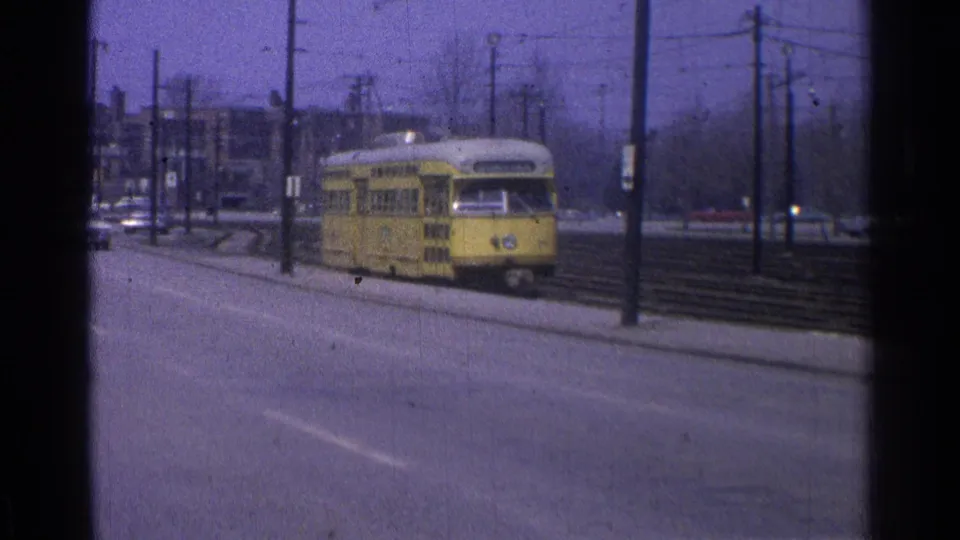 1965 Ohio home-movie frame showing a yellow trolley closer to camera, with road lanes, poles, tracks, and background buildings.