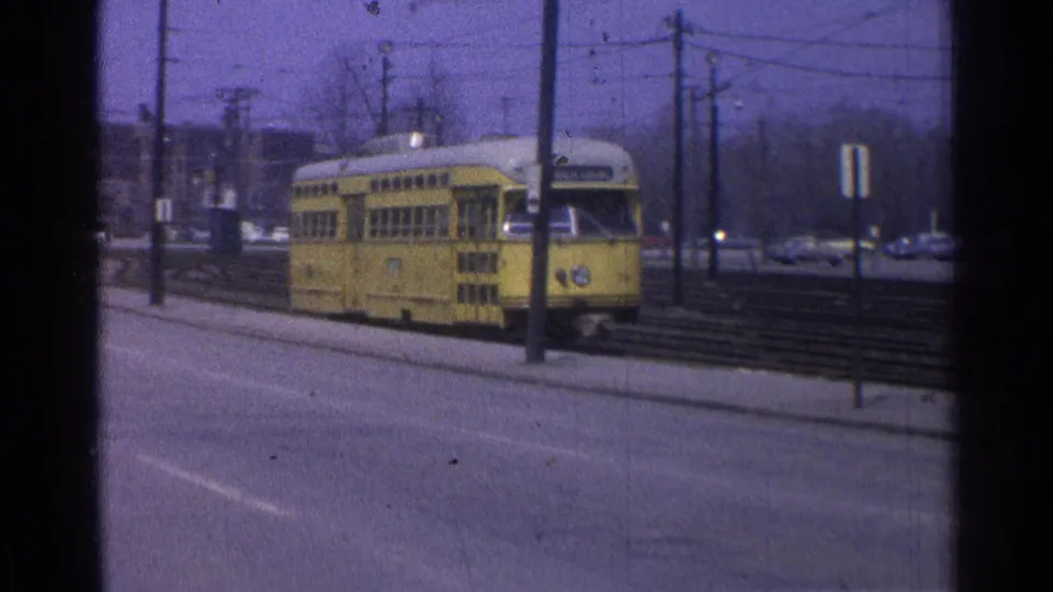 1965 Ohio frame of a yellow trolley moving through a street corridor with rails, utility poles, parked cars, and hazy winter trees.