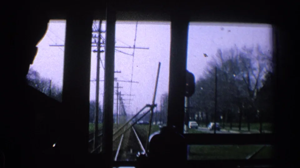 1965 Ohio home-movie frame looking through transit windows past a silhouetted rider toward poles, trees and cars on the road.