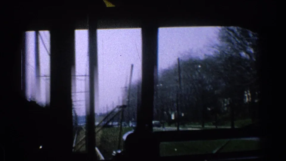 8mm interior view from the trolley or streetcar, showing window frames, overhead wires, bare trees and cars in cloudy daylight.