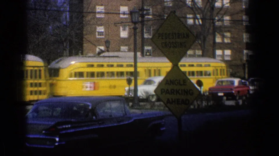 A yellow electric train passes a suburban Ohio street in 1965, with classic cars, road signs, trees, and brick apartments visible.