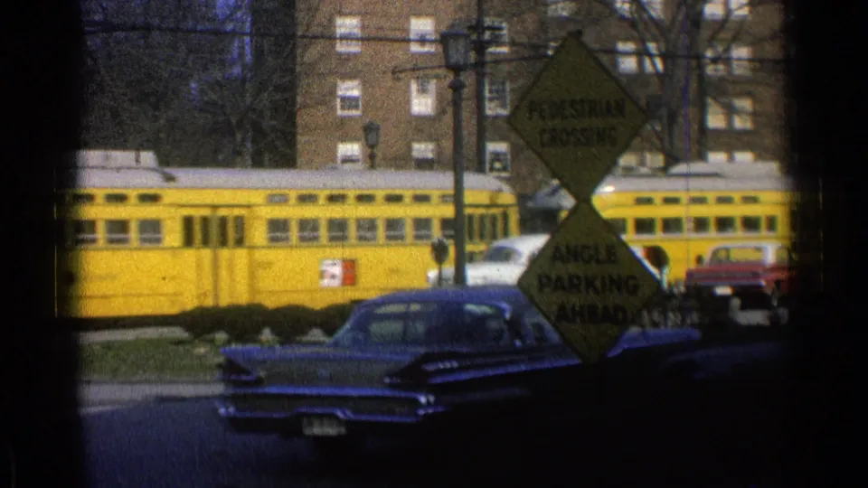 The yellow train continues through the suburban street scene, passing cars, road signs, trees, and a brick apartment building.