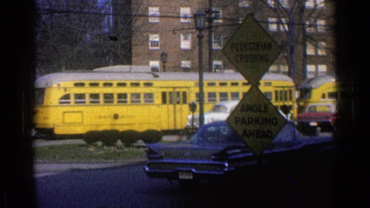 Yellow rail car passes behind parked cars and road signs near brick apartment buildings.