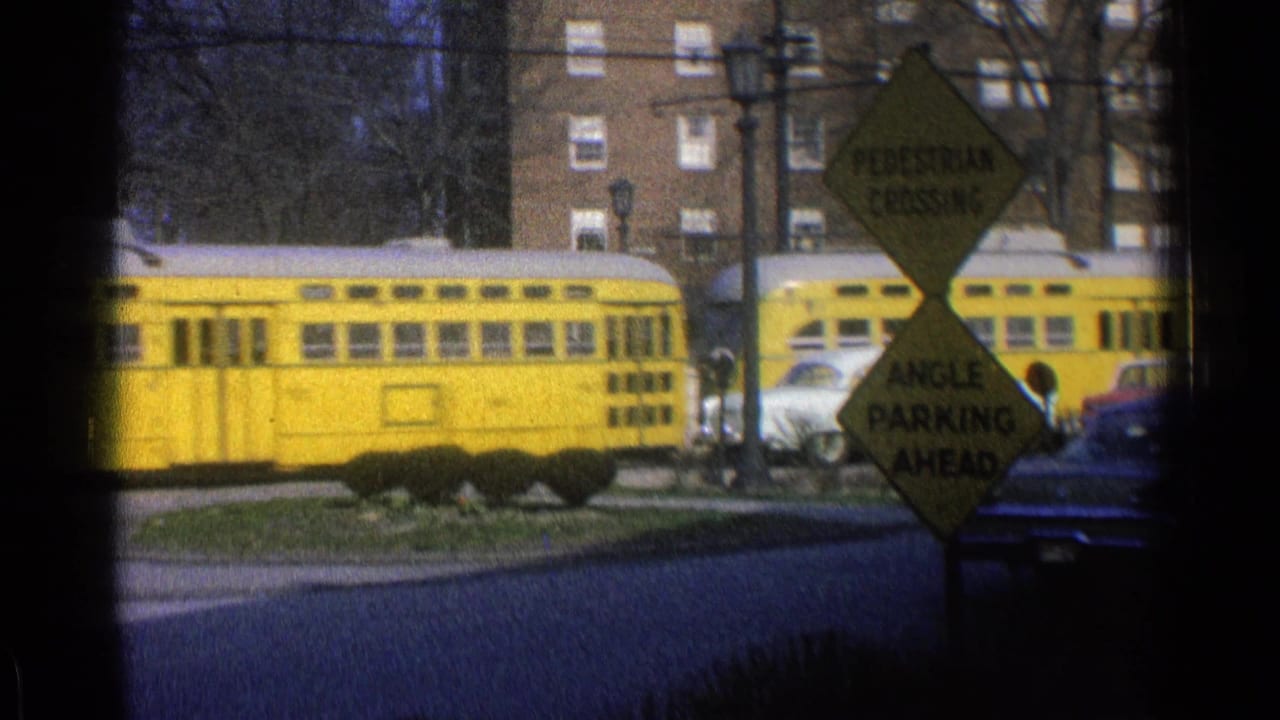 Yellow rail car moves past a street corner with road signs and parked cars.