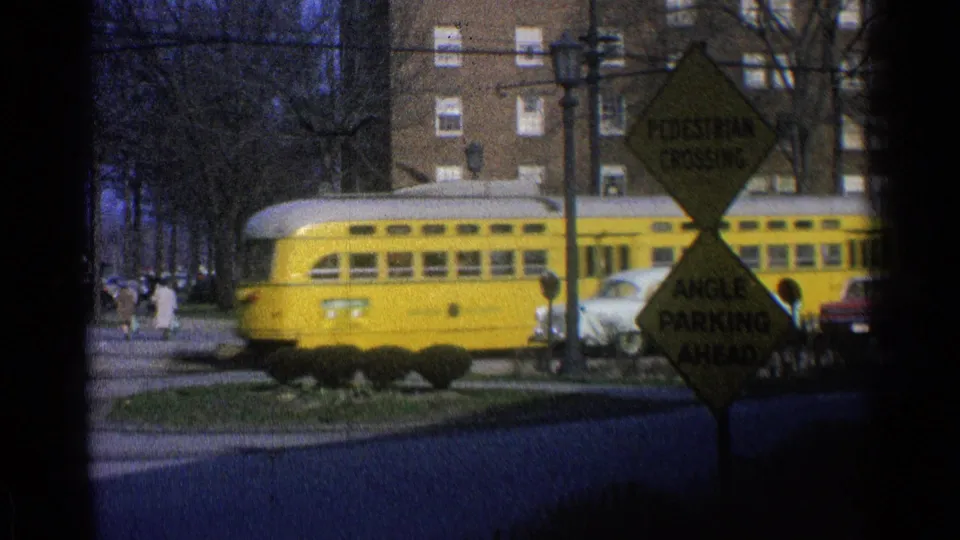 A yellow commuter rail car crosses the frame in 1965 Ohio, with street signs, parked cars, and brick apartment blocks visible behind it.