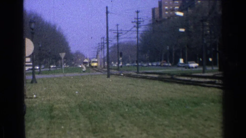 1965 Ohio 8mm frame showing a distant yellow trolley approaching along tracks beside grass, utility poles, bare trees, cars, and a brick building.
