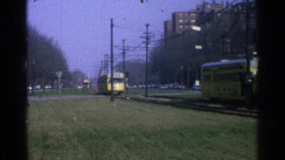 1965 Ohio 8mm view of two yellow trolleys moving in opposite directions on city tracks beside grass, utility poles, street signs, and distant cars.
