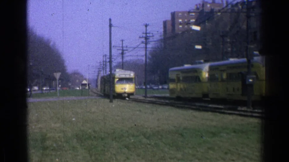 1965 Ohio frame showing two yellow trolleys passing on parallel city tracks, with grass, overhead wires, poles, street signs, cars, and brick buildings visible.