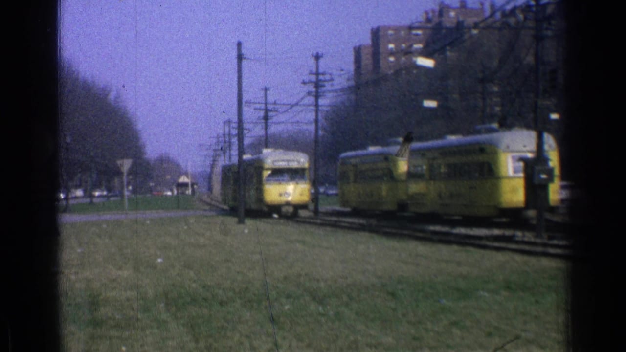 1965 Ohio 8mm frame showing two yellow trolleys on parallel tracks with utility poles, grass, trees, and apartment buildings in the background.