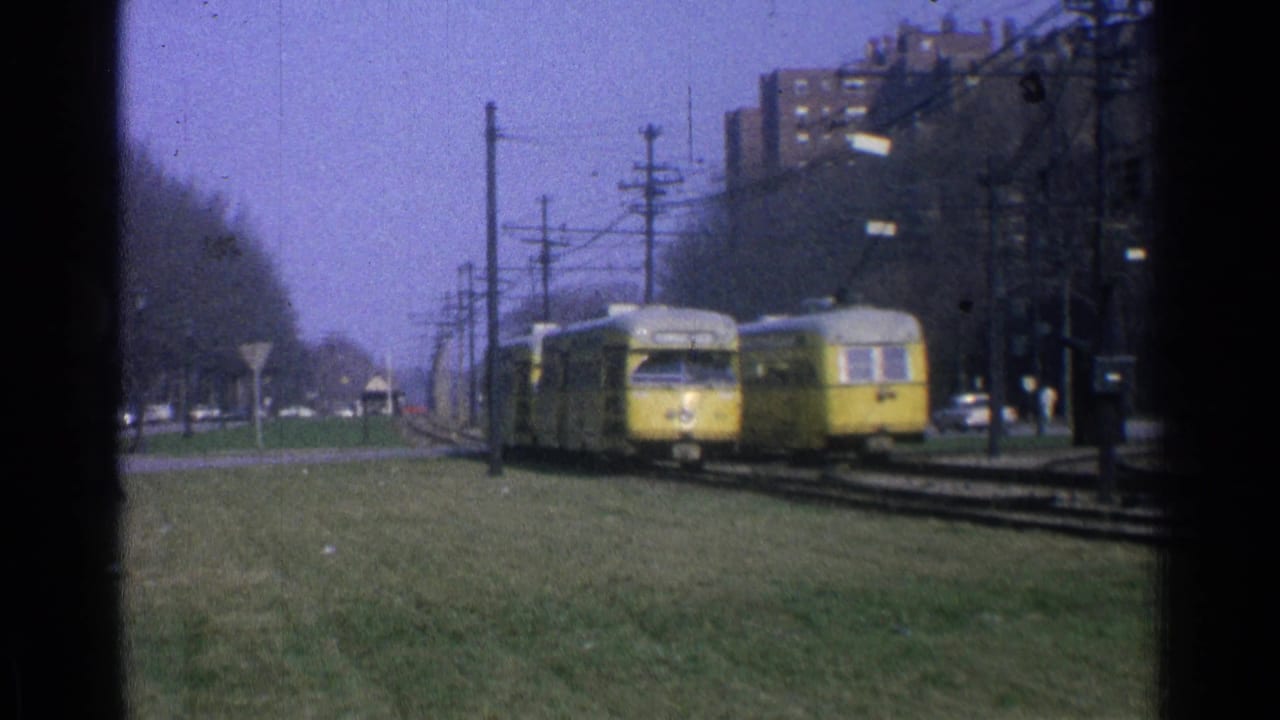 1965 Ohio trolley scene with two yellow cars passing on a grassy right-of-way under overhead power lines near brick apartments.