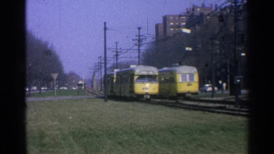 1965 Ohio trolley scene with two yellow cars passing on a grassy right-of-way under overhead power lines near brick apartments.