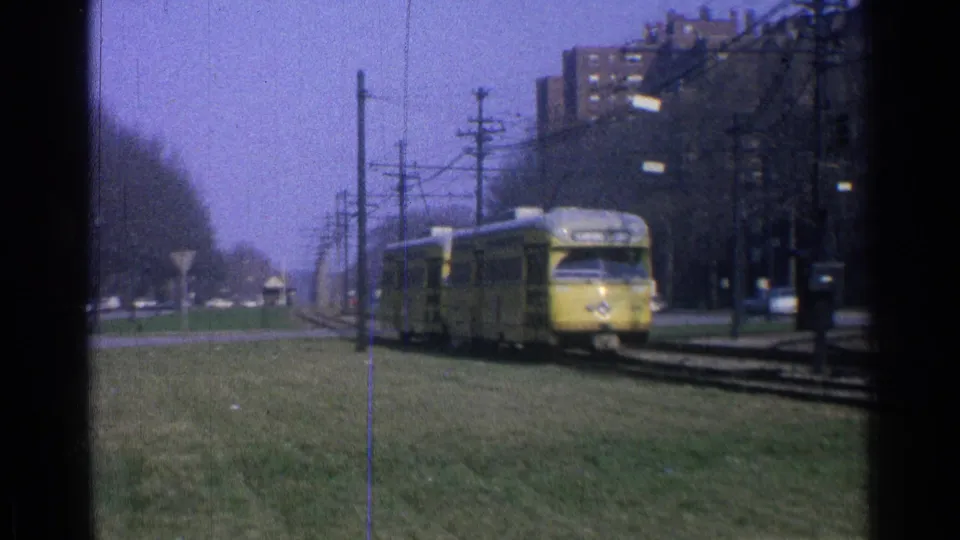 Yellow 1960s trolley car advances along an Ohio track corridor, with grass, overhead lines, trees, and apartment buildings visible.