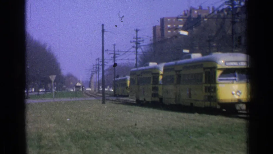 1965 Ohio 8mm frame with a yellow trolley passing close at right, another trolley behind it, and overhead wires along the grassy corridor.