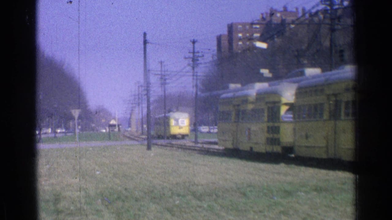 1965 Ohio home-movie frame showing a yellow trolley near city tracks, with grass, overhead wires, cars, and apartment buildings beyond.