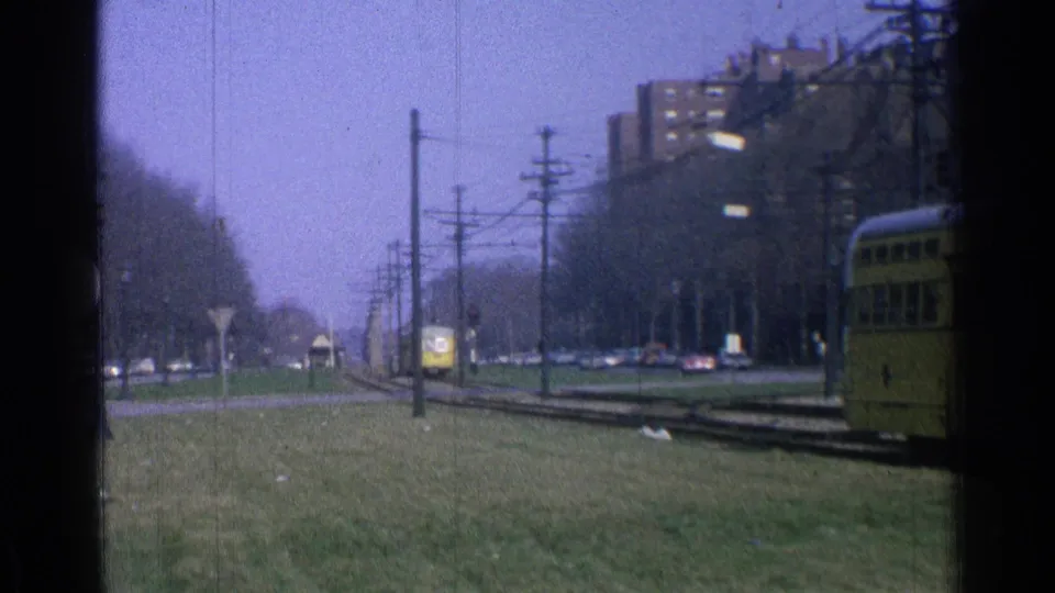 1965 Ohio home-movie view of a distant yellow trolley on tracks, with a cropped trolley at right, grass, wires, cars, and apartments.