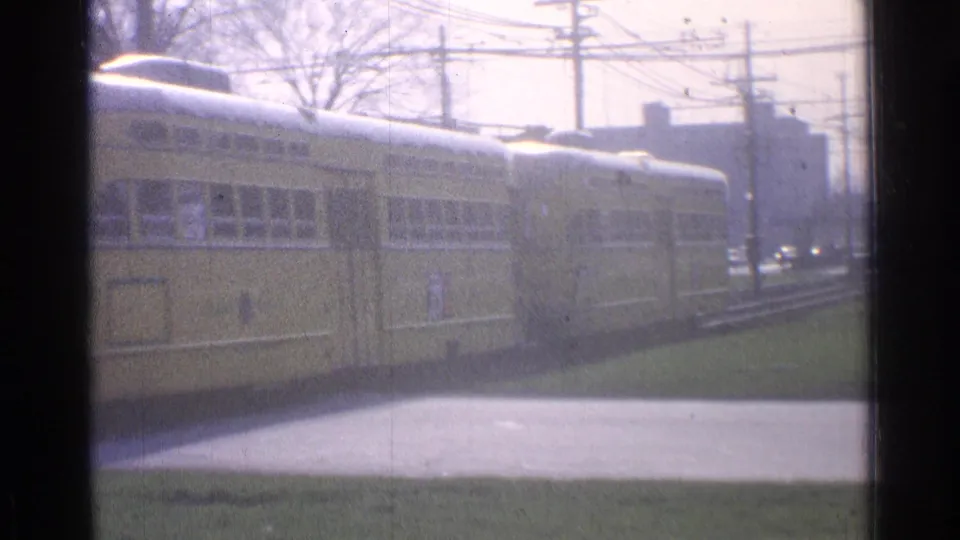 Close 1965 Ohio frame of a yellow trolley passing the camera, softened by haze, with windows, wires, grass, and buildings behind.