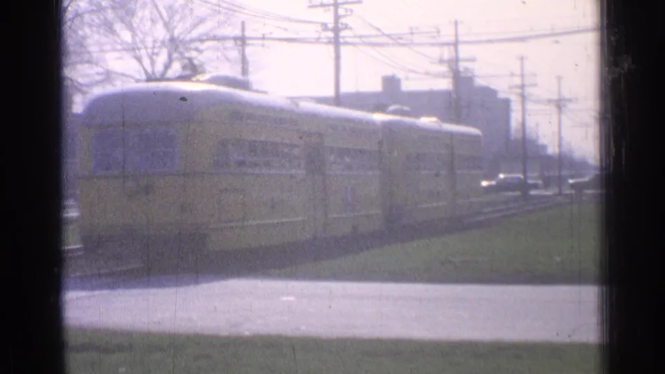 1965 Ohio 8mm frame showing a yellow trolley car on urban tracks, seen through dark window framing with utility lines and buildings behind it.