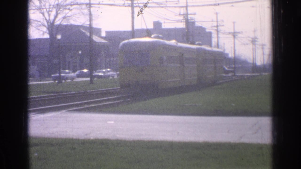 The trolley continues along Ohio city tracks in soft 8mm focus, with grass, roadway, cars, utility poles, and low buildings in the distance.