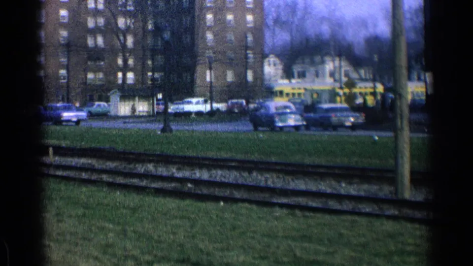 1965 Ohio street view from 8mm footage, showing rail tracks in the foreground, parked cars, small houses, and brick apartment buildings.