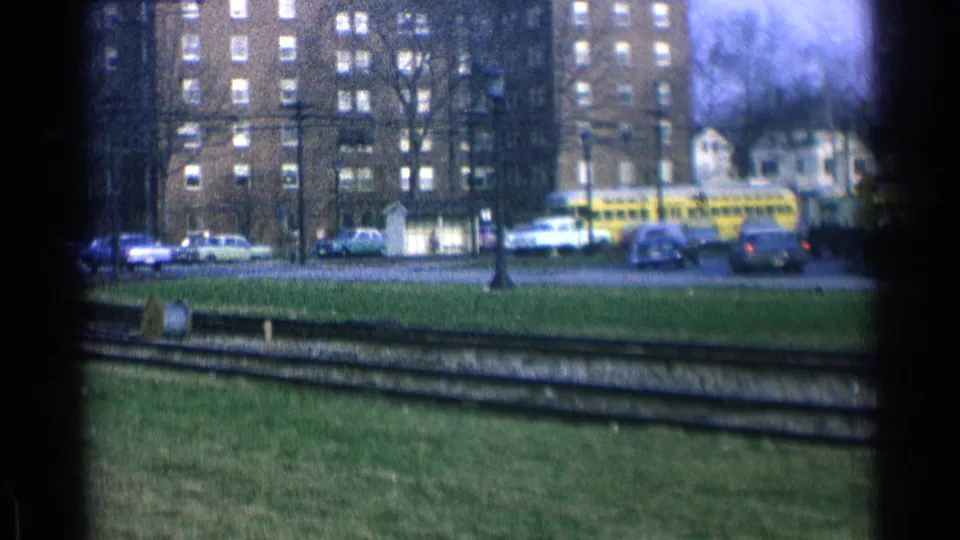 A yellow trolley appears across the street in this 1965 Ohio home-movie frame, with rail tracks, grass, cars, trees, and brick apartments visible.