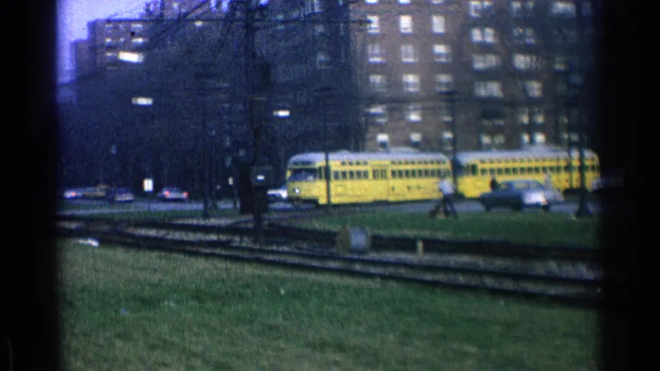 A two-car yellow trolley moves through the Ohio street scene, with curved tracks, grass, cars, buildings, and a small distant pedestrian.
