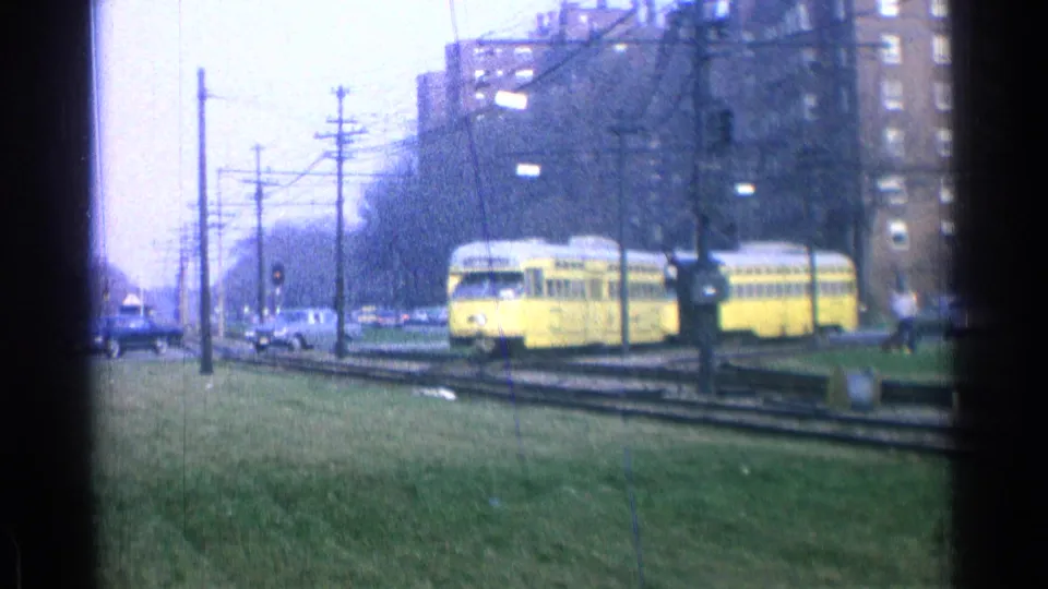 The yellow trolley rounds or crosses the track area in the 1965 Ohio scene, with utility poles, cars, grass, and apartment blocks visible.