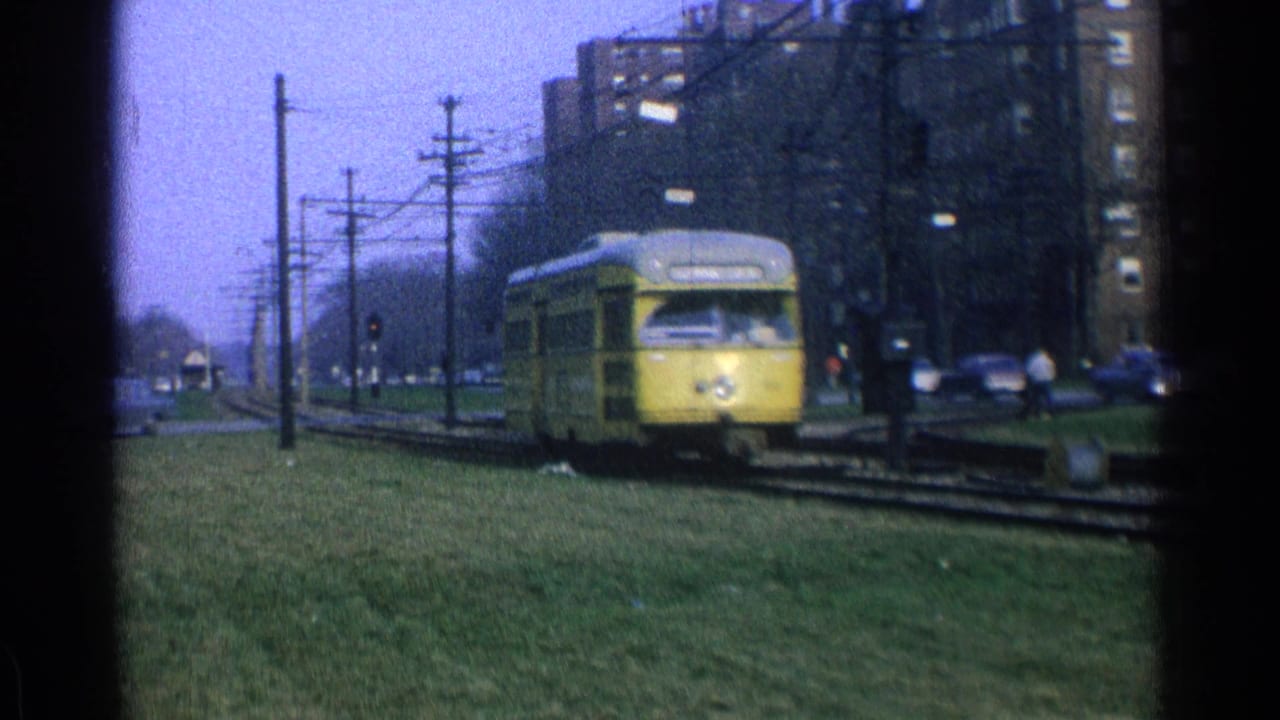 1965 Ohio 8mm frame of a yellow trolley coming toward the camera on urban tracks with cars, wires, and apartment buildings nearby.