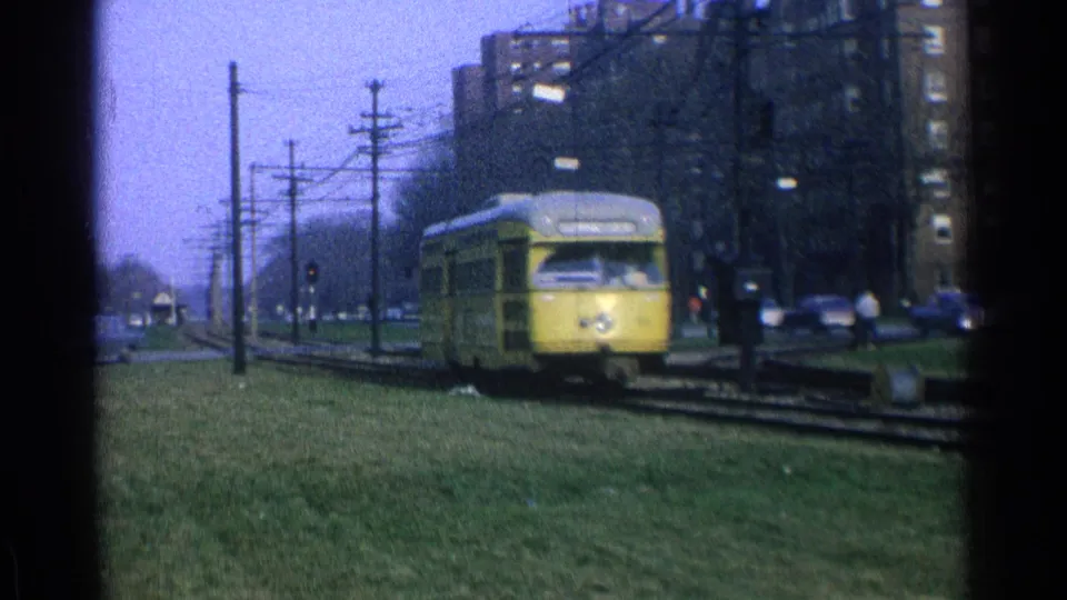 1965 Ohio 8mm frame of a yellow trolley coming toward the camera on urban tracks with cars, wires, and apartment buildings nearby.