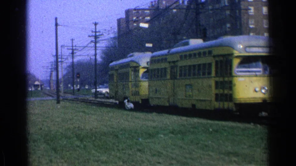 1965 Ohio 8mm frame showing yellow trolley cars passing near the camera with tracks, grass, wires, and apartment buildings visible.