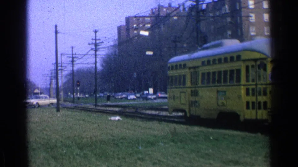 1965 Ohio 8mm frame of the rear of a yellow trolley heading away along tracks beside grass, cars, utility poles, and apartment buildings.