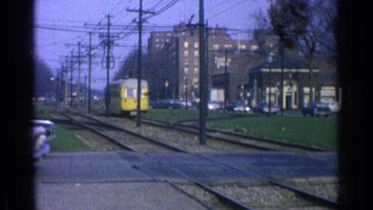 Distant yellow trolley near a station-like structure beside tracks, cars, and overhead wires.