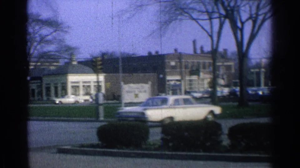 A white sedan passes through an Ohio city street scene, with a low brick building, bare trees, parked cars, and shrubs visible.